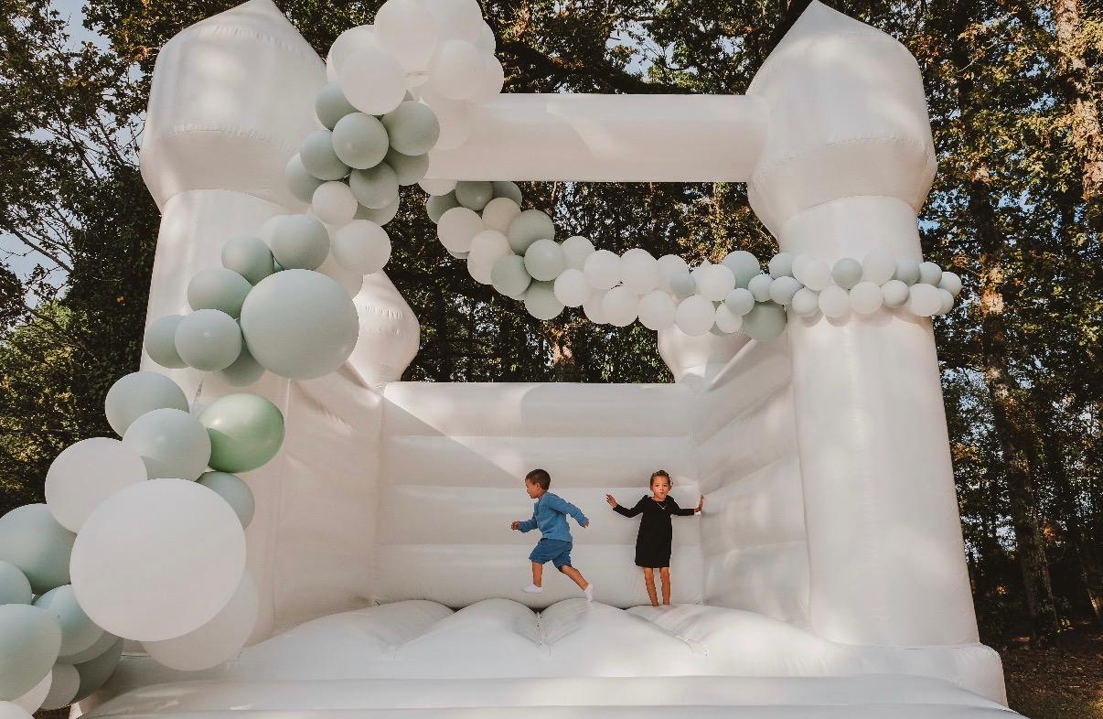 Château gonflable entièrement blanc décoré d'une arche de ballons blancs et vert sauge, lors d'un mariage