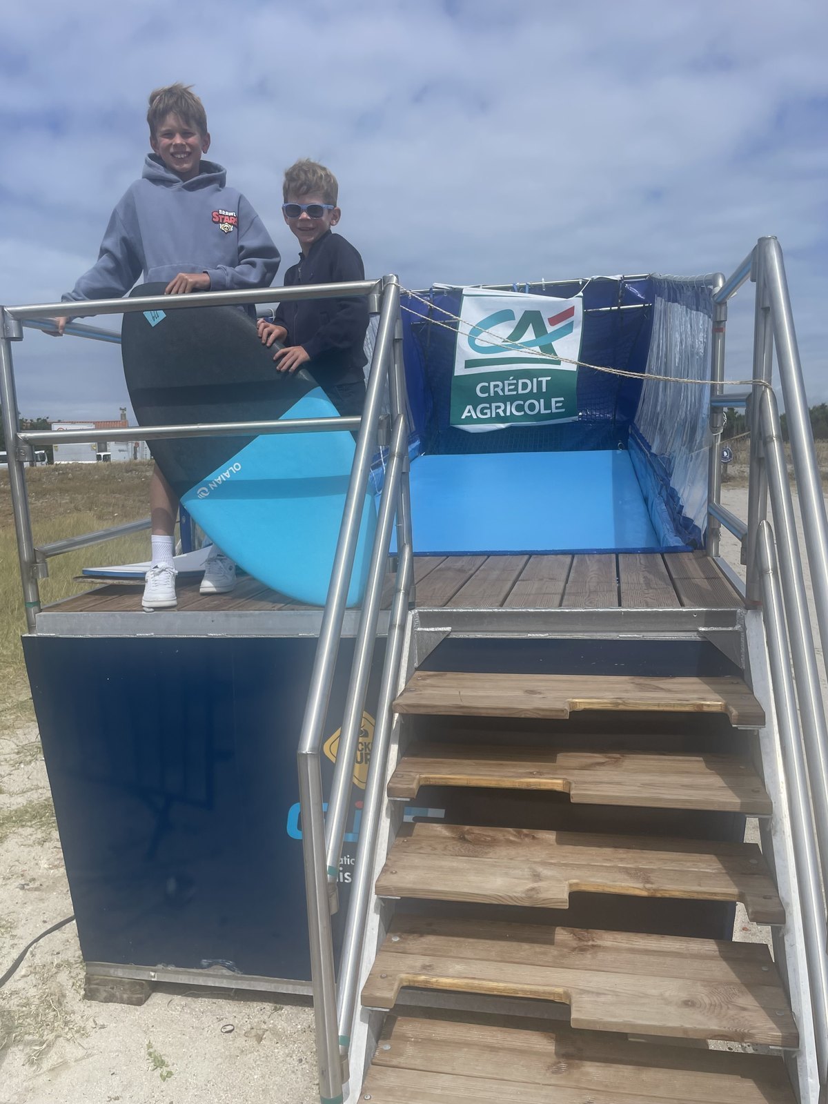Deux enfants tiennent leurs planches sur la passerelle du simulateur de surf mobile